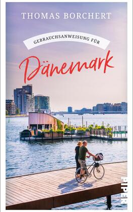 Gebrauchsanweisung für Dänemark Das Buchcover zeigt einen malerischen Ausblick auf einen Hafen mit modernen Gebäuden im Hintergrund. Im Vordergrund sind ein Paar und ein Radfahrer auf einem Holzsteg abgebildet. Der Himmel ist klar und in sanften Farbtönen, die eine entspannte Atmosphäre erzeugen. Der Titel „Gebrauchsanweisung für Dänemark“ ist in großen, roten Buchstaben platziert, während der Autor „Thomas Borchert“ oben in eleganter Schrift erscheint.