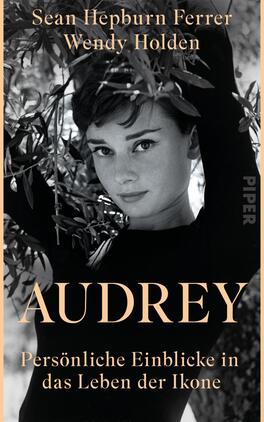 Audrey Das Buchcover zeigt eine Schwarz-Weiß-Fotografie von einer Frau mit einem sanften Lächeln, umgeben von grünen Olivenzweigen. Sie trägt einen schwarzen Rollkragenpullover und blickt direkt in die Kamera. Der Titel „AUDREY“ ist in großen, goldenen Buchstaben in der Mitte platziert, darunter steht der Untertitel „Persönliche Einblicke in das Leben der Ikone“ in kleinerer, heller Schrift. Die beiden Autoren, Sean Hepburn Ferrer und Wendy Holden, sind oben in eleganter, weißer Schrift angeführt. Die Atmosphäre ist nostalgisch und stilvoll.