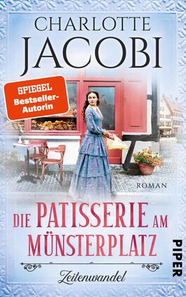 Die Patisserie am Münsterplatz – Zeitenwandel Das Buchcover zeigt eine historische Szene mit einer weiblichen Figur in einem eleganten, blauen Kleid, die vor einem charmanten, roten Patisserie-Geschäft steht. Die Atmosphäre strahlt Nostalgie und Gastronomie aus. Der Titel „Die Patisserie am Münsterplatz“ ist in geschwungener Schrift in der unteren Mitte platziert, während der Autorennamen „Charlotte Jacobi“ oben hervorgehoben wird. Ein orangefarbener Hinweis „SPIEGEL Bestseller-Autorin“ zieht die Aufmerksamkeit auf sich. Der Hintergrund ist hell und freundlich gestaltet.