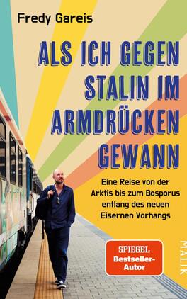 Als ich gegen Stalin im Armdrücken gewann Das Buchcover zeigt einen bärtigen Mann in einem blauen Anzug, der an einer Bahnstation steht. Er blickt nachdenklich in die Ferne. Im Hintergrund verläuft ein Zug, und der Himmel ist in sanften Farben mit gelb, orange, grün und blau abgebildet, die eine dynamische Atmosphäre schaffen. Der Titel „ALS ICH GEGEN STALIN IM ARMDRÜCKEN GEWANN“ steht in großen, fetten weißen Buchstaben, während der Untertitel „Eine Reise von der Arktis bis zum Bosporus entlang des neuen Eisernen Vorhangs“ in etwas kleinerer Schrift darunter platziert ist.