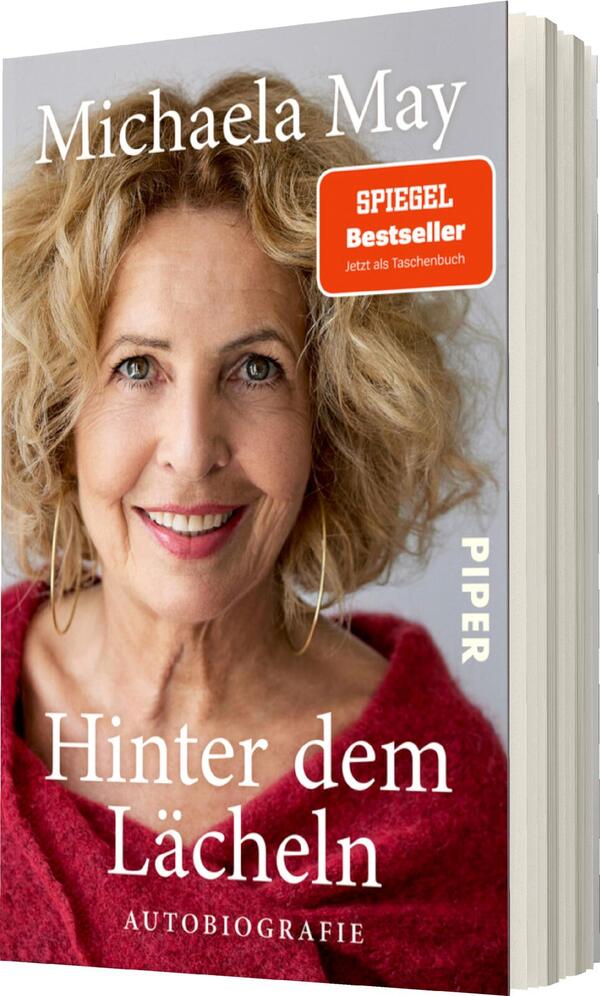 Das Buchcover zeigt das Portrait von Michaela May, die freundlich lächelnd in die Kamera schaut. Ihr volles, lockiges Haar umrahmt das Gesicht. Der Hintergrund ist hell und unauffällig, während ihr rotes Oberteil einen warmen Farbton ausstrahlt. Der Titel „Hinter dem Lächeln“ ist in auffälliger, weißer Schrift platziert, darunter steht „AUTOBIOGRAFIE“ in kleinerer Schrift. Ein rotes Etikett informiert, dass das Buch ein „SPIEGEL Bestseller“ ist und jetzt als Taschenbuch erhältlich ist.