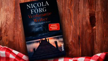 Das Buch 'Verdammte Weiber' von Nicola Förg liegt auf einem Holztisch. Auf dem Cover ist ein Steg, der in einen stürmischen Himmel mit Blitzen ragt. Daneben liegt ein rotes kariertes Tuch.