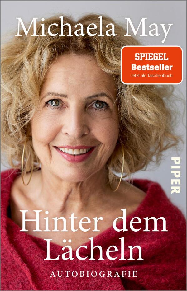 Das Buchcover zeigt ein Portrait von Michaela May mit lockigem, blonden Haar und einem warmen Lächeln. Der Hintergrund ist hell und freundlich. Oben links steht der Name „Michaela May“ in großer, eleganter Schrift. Darunter ist der Titel „Hinter dem Lächeln“ in auffälliger roter Schrift, gefolgt von dem Zusatz „AUTOBIOGRAPHIE“ in kleinerer Schrift. Oben rechts befindet sich ein rotes Label mit der Aufschrift „SPIEGEL Bestseller“, ergänzt durch den Hinweis „Jetzt als Taschenbuch“.