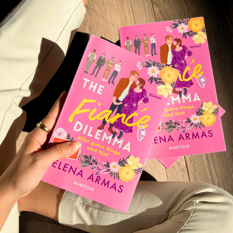 „The Fiancé Dilemma“ von Elena Armas wird in einer Hand gehalten. Ein zweites Exemplar liegt auf dem Boden.