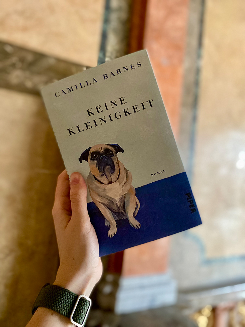 Eine Hand hält ein Buchcover. Der Titel lautet 'Keine Kleinigkeit' von Camilla Barnes. Auf dem Cover ist ein sitzender Hund abgebildet.