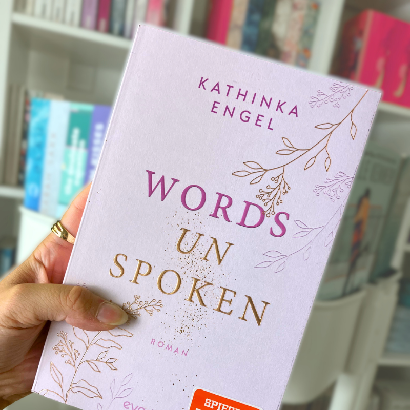 „Words Unspoken“ von Kathinka Engel wird vor einem Bücherregal gehalten.