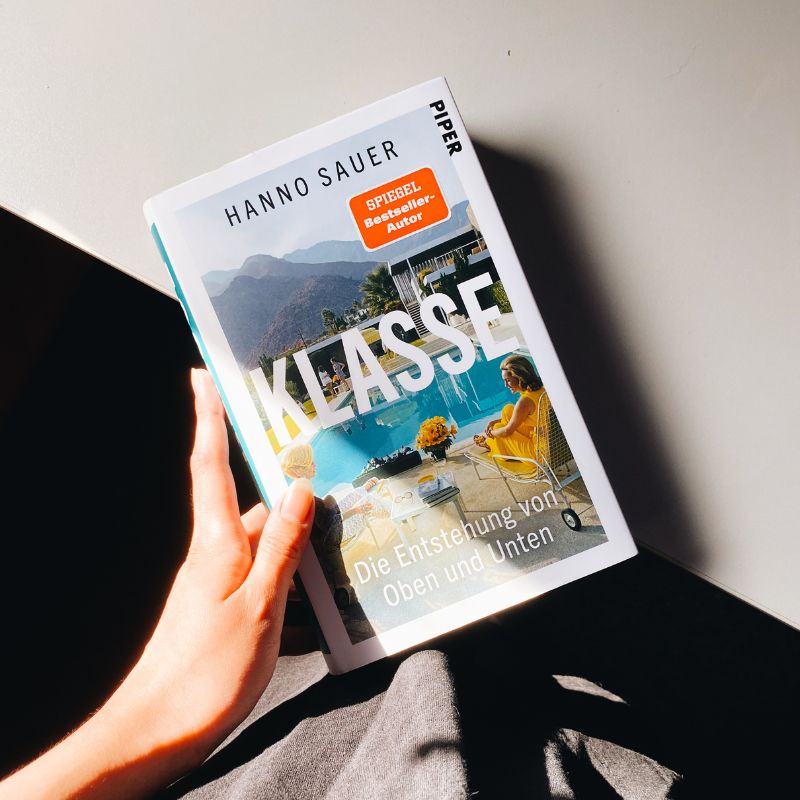 "Klasse" von Hanno Sauer liegt zur Hälfte auf einem weißen Tisch und wird von einer Hand gehalten.